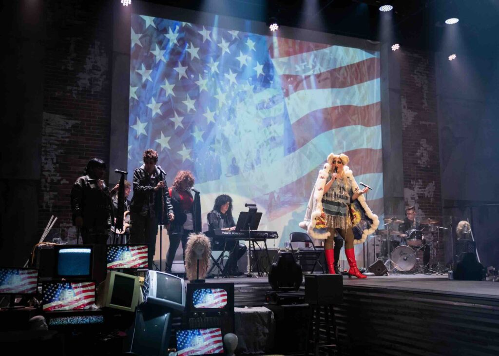 A stage production of "Hedwig and the Angry Inch" with performers and musicians on stage. A large American flag is projected on a screen behind them. The lead performer, likely Hedwig, stands to the right wearing a flamboyant white costume with fringe and fur trim, and red boots. Band members in dark clothing are positioned around instruments including a keyboard and drums. The foreground shows old TV sets decorated with small American flags. The setting has brick walls and theatrical lighting.